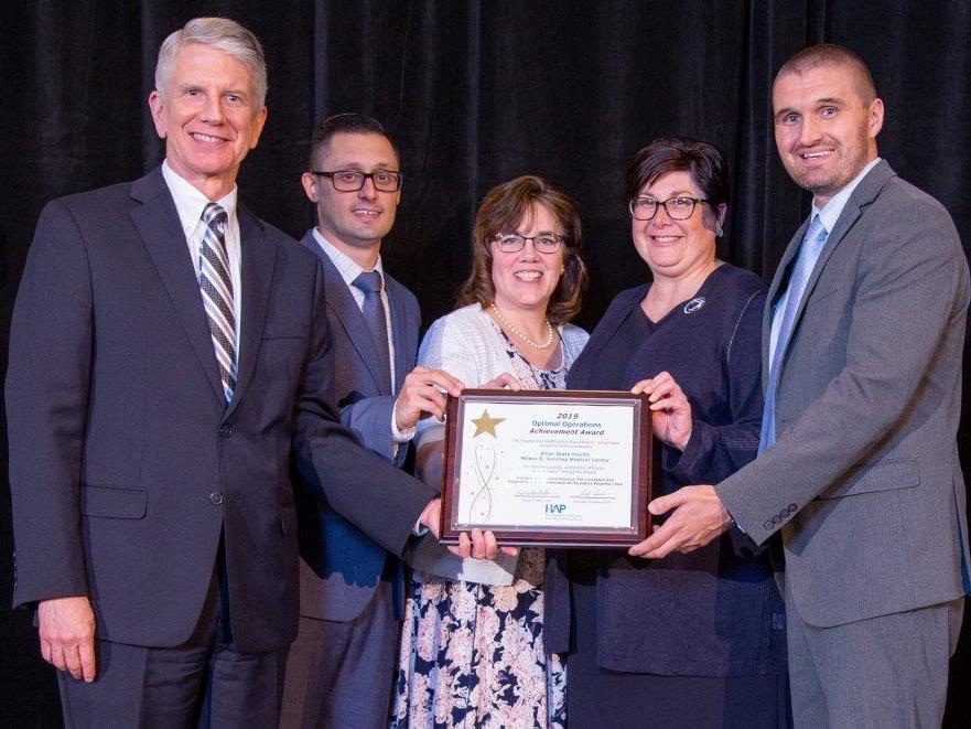 Five people – two women and three men, pose for a photo, wearing semi-formal/business attire, each with a hand on a framed certificate that reads “2019 Optimal Operations Achievement Award.”