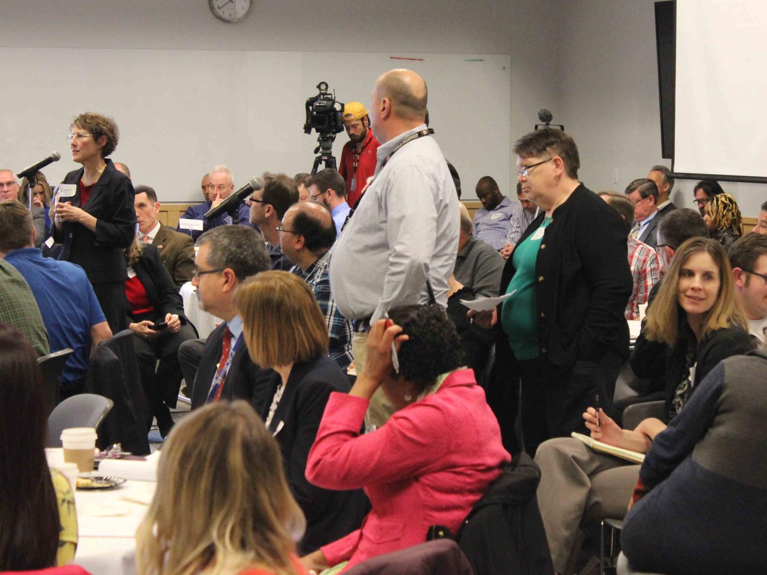 Several people are seated, all facing the same direction. A few people are standing at microphones placed in the middle of the crowd.
