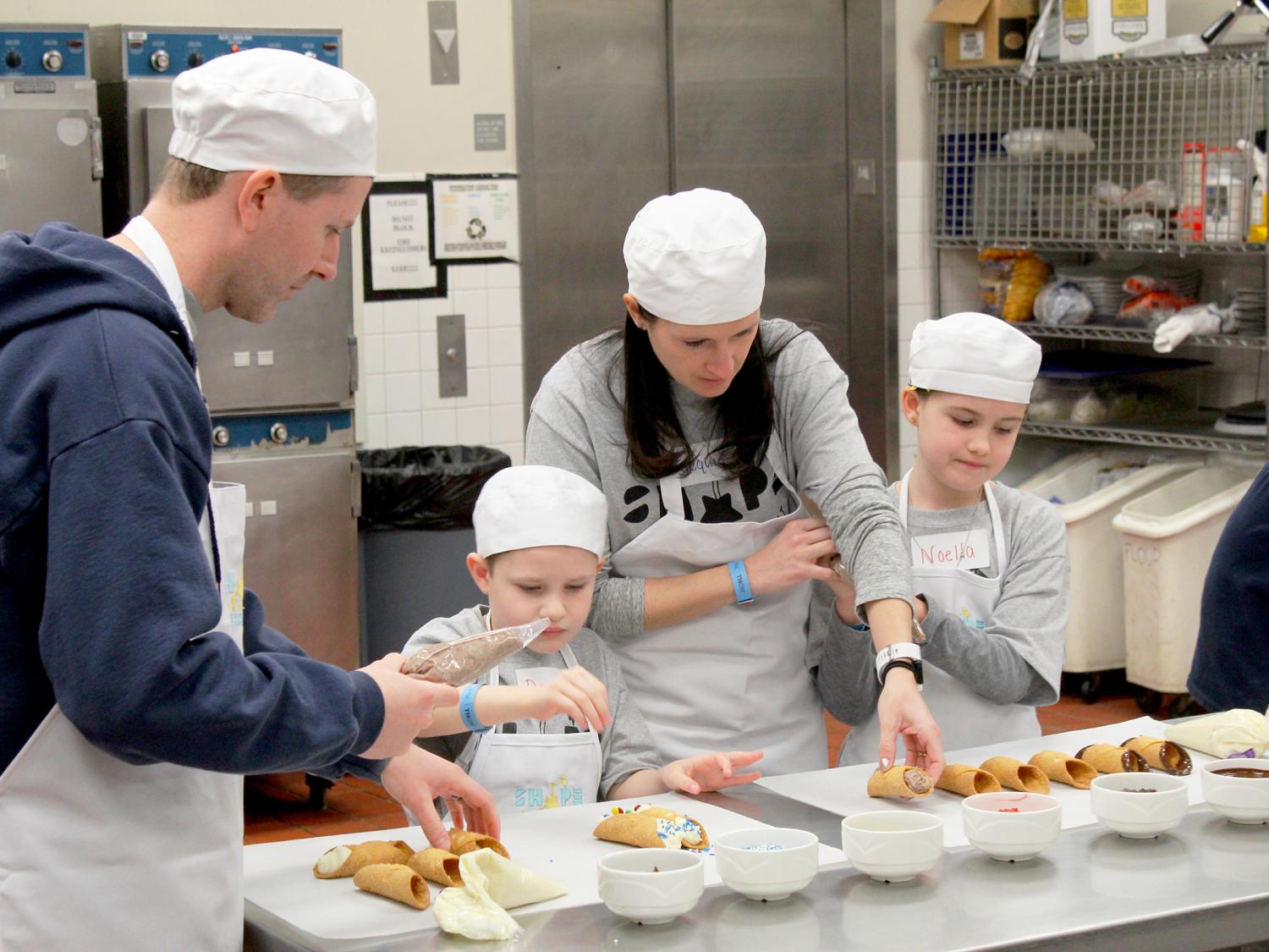 A man, a woman, and two children make cannoli in a restaurant kitchen