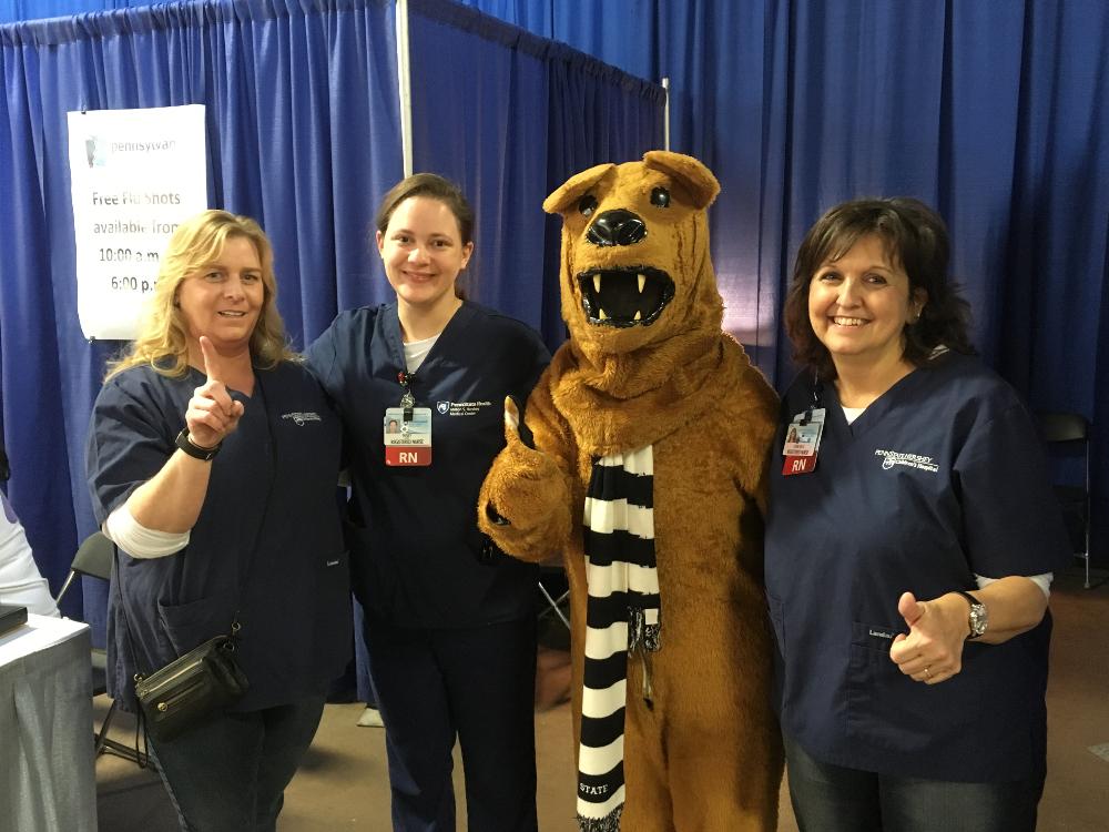 Three Penn State Health nurses pose with the Nittany Lion, in front of a blue pipe-and-drape backdrop.