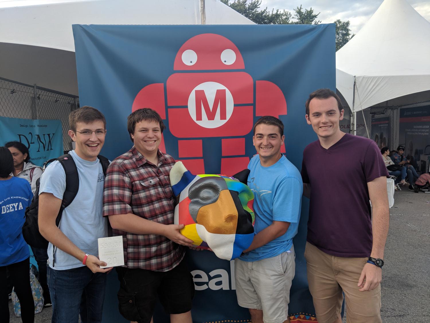 Four men stand in front of a banner and hold a 3D printed Nittany Lion Shrine Head