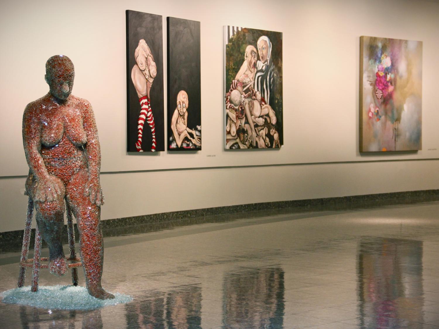 On the left foreground, a life-sized sculpture of a seated female drips broken glass. Behind her, three paintings depicting human-like, yet still somewhat alien, figures against dark backgrounds.