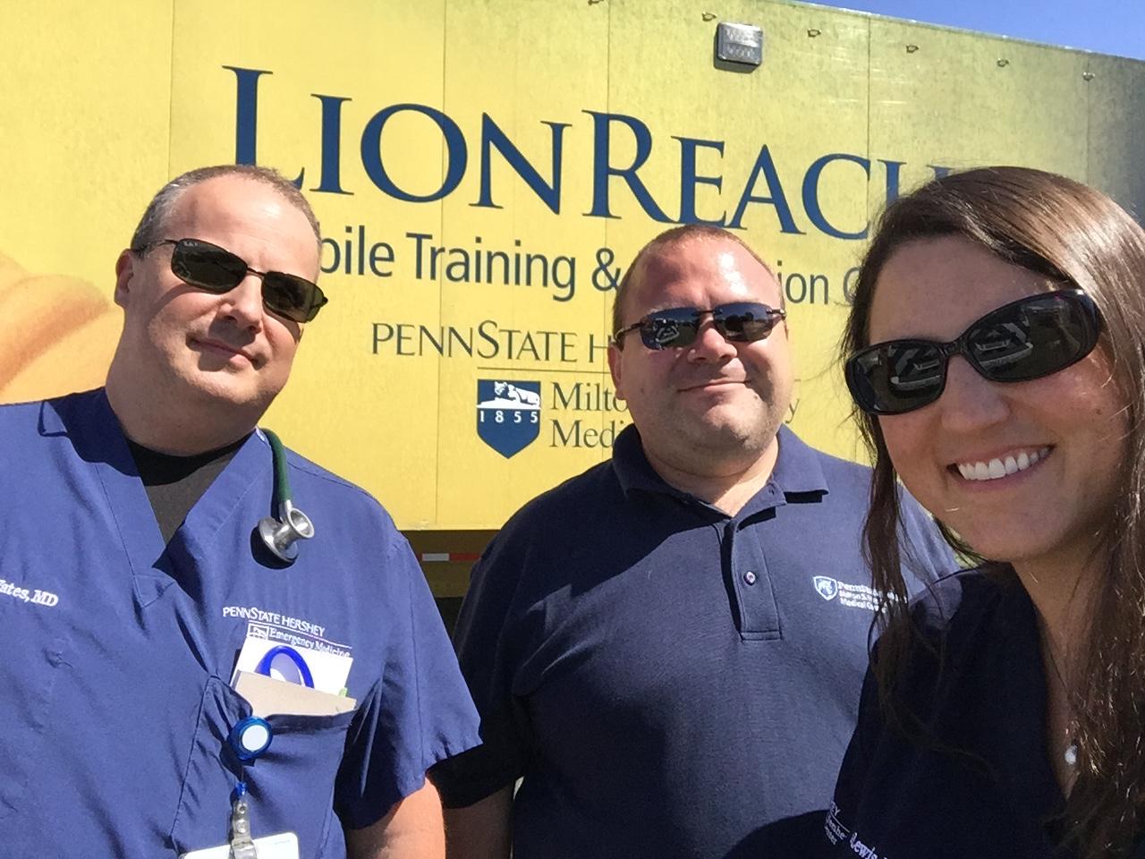 Three people – two men and one woman – pose for a photo in front of a large trailer that has the word “LionReach” on it as well as the Penn State Health Milton S. Hershey Medical Center logo.