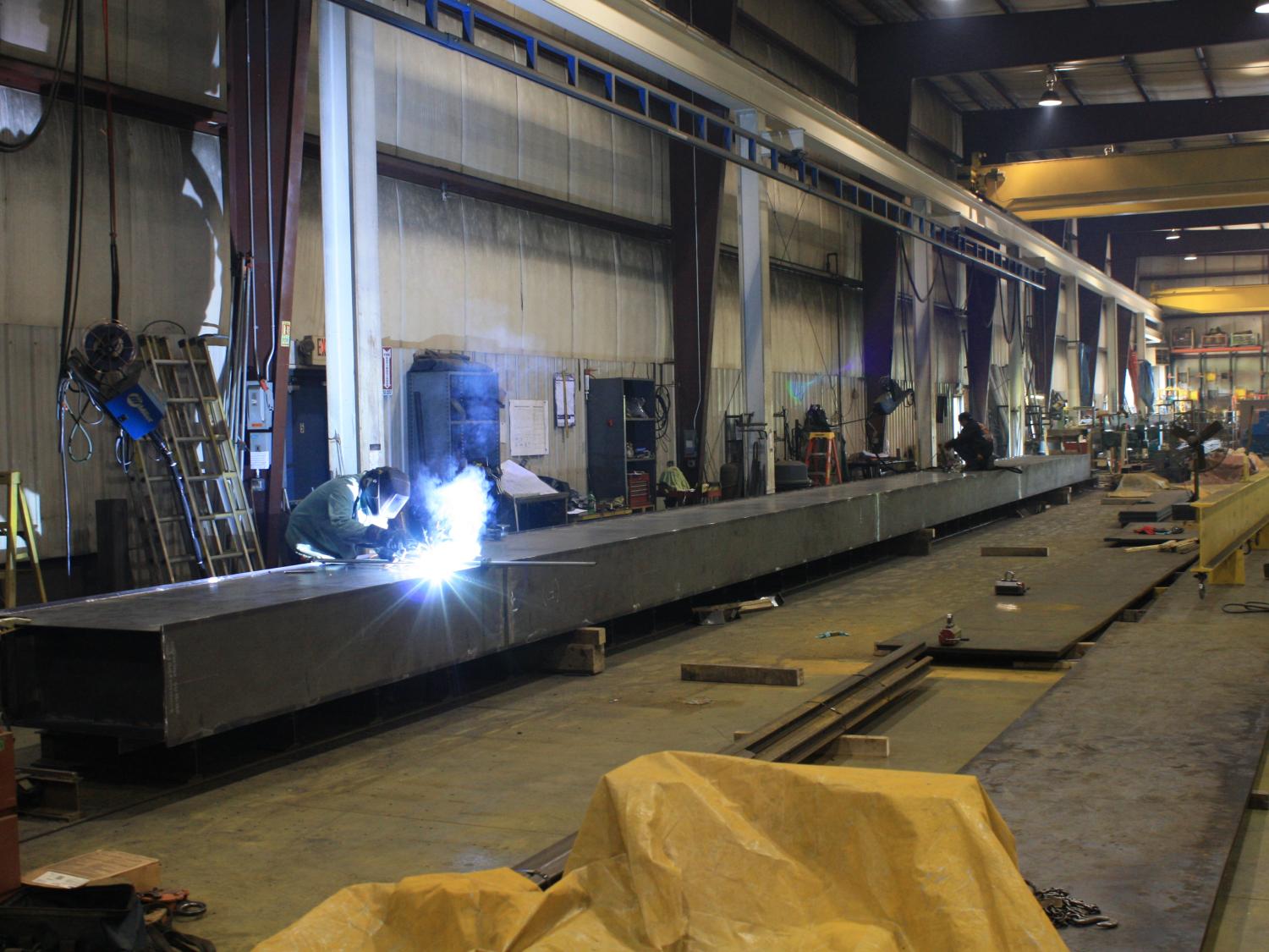 Factory employees work on a metal beam. One employee is welding the metal. 
