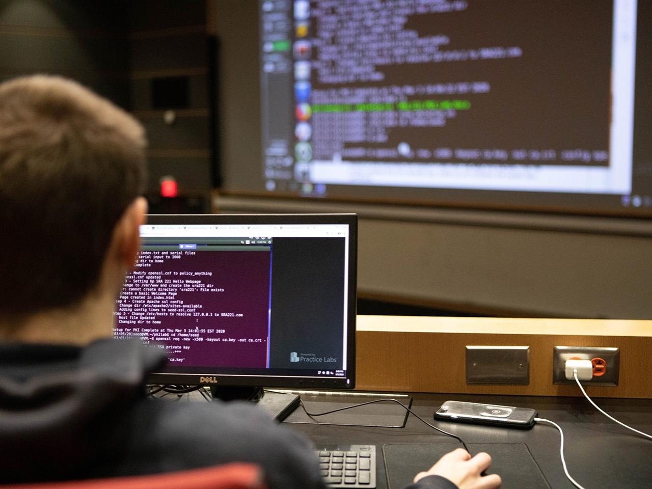 A male student codes on a computer screen while a teacher instructs on a large screen at the front of the classroom.