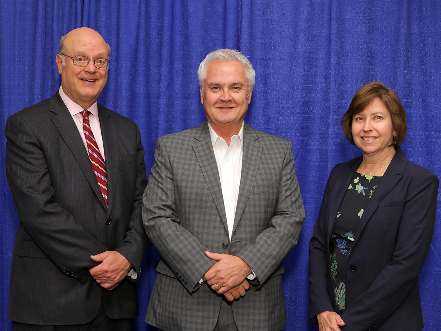 Three people – two men and a woman – wearing professional attire pose for a photo in front of a blue curtain.