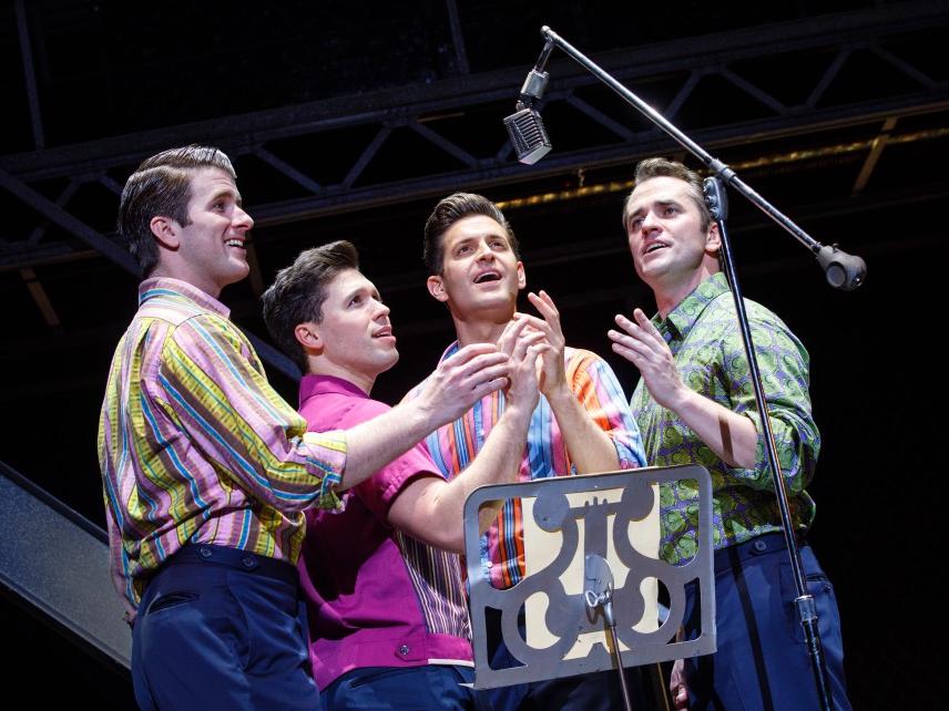Four men wearing 1960s-era casual clothing crowd around a suspended micrphone to sing.