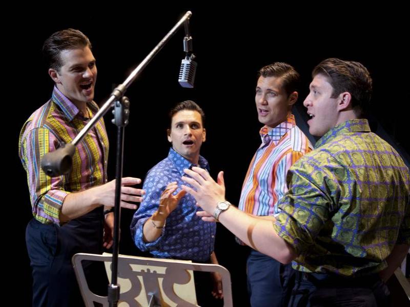 The cast of "Jersey Boys," gathered around a microphone, sings a song.