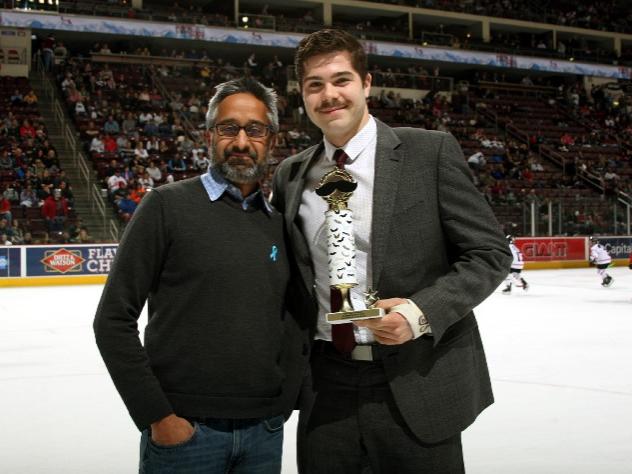 Two men pose for a photo, looking at the camera. The man on the right holds a small trophy. They are standing on an ice rink, where some young hockey players and fans are seen in the background.