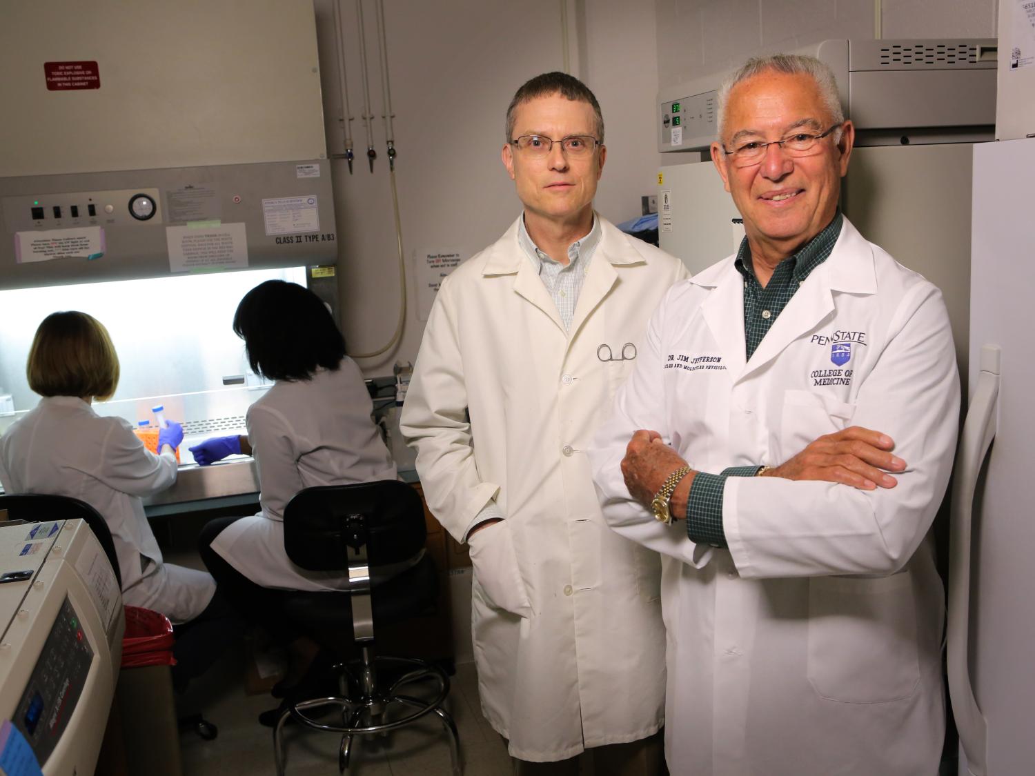 Two men in white coats stand at right, looking toward the camera. In the background at left, two women face away from the camera, working under a laboratory hood.