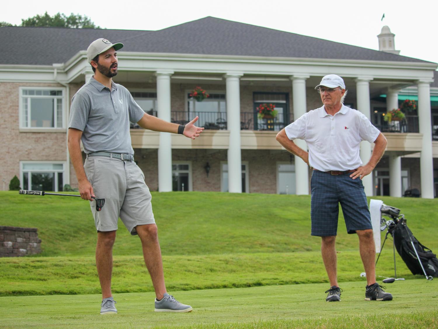 Two men stand on a golf course, one is talking. 