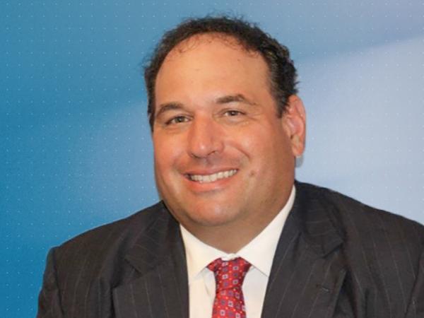 Headshot of Jonathan Kukta, Penn State Schuylkill's director of business services, wearing a gray suit, white shirt, and red tie, and standing in front of a blue background