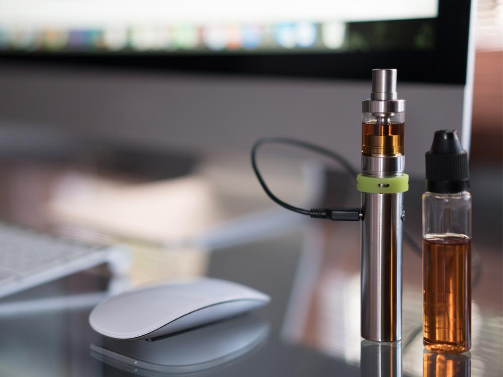 A close-up of a vaping device and a small vial of vaping liquid sitting on a counter. A computer keyboard, monitor and mouse sit nearby, slightly out of focus.