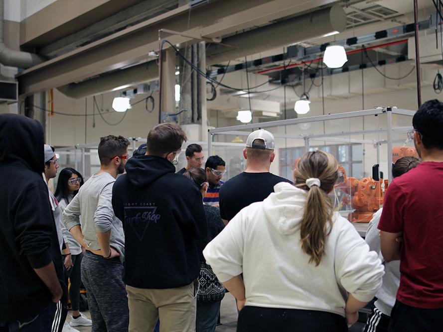  A large group of students stand around a robot in a glass encasement.