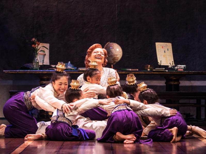 A woman sitting on the floor is surrounded by girls in traditional Siam clothing who smile and give her hugs.