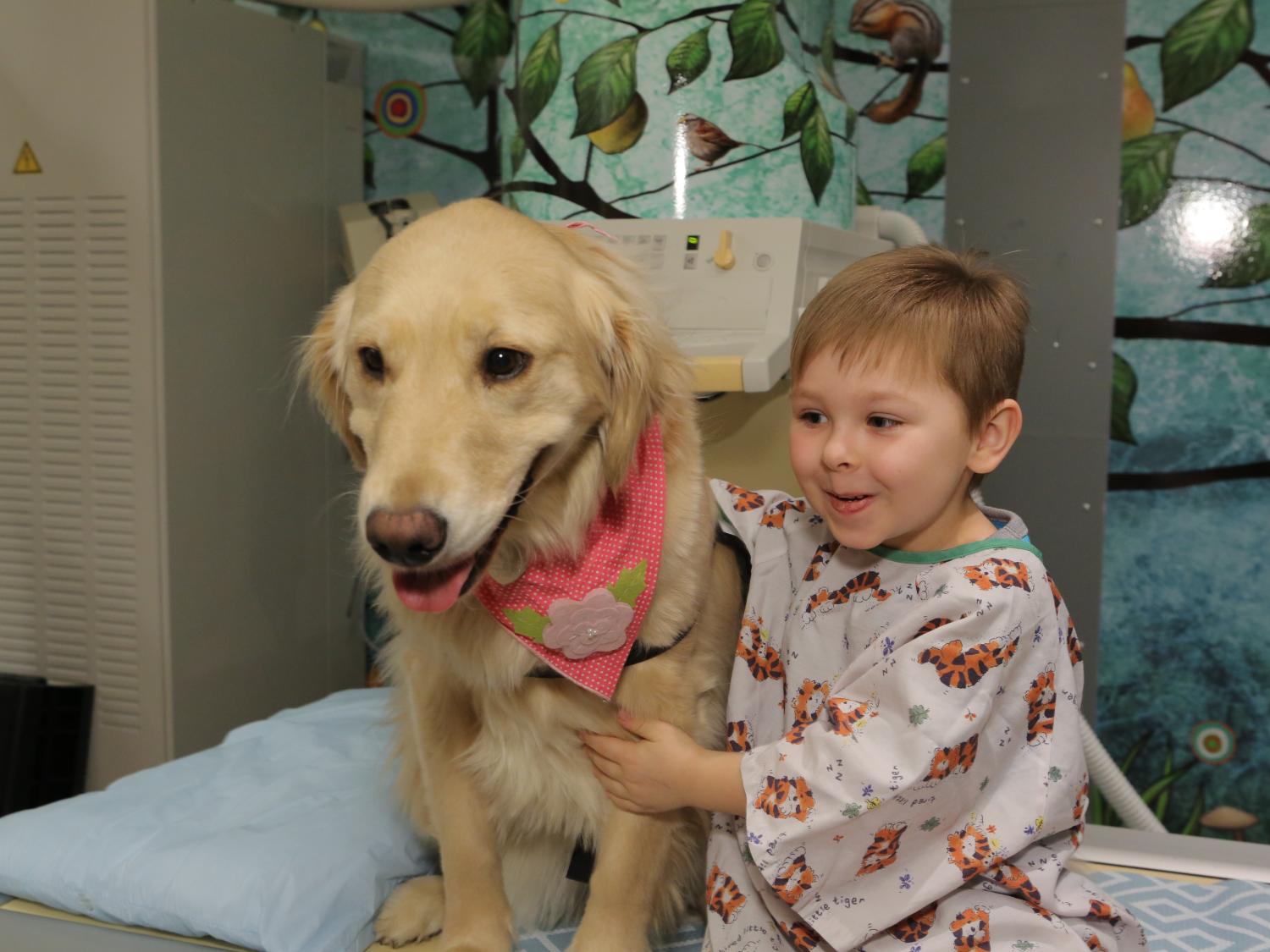 A boy in a hospital gown sits alongside a golden retriever. The boy is smiling; the dog is wearing a pink scarf with a flower around its neck.