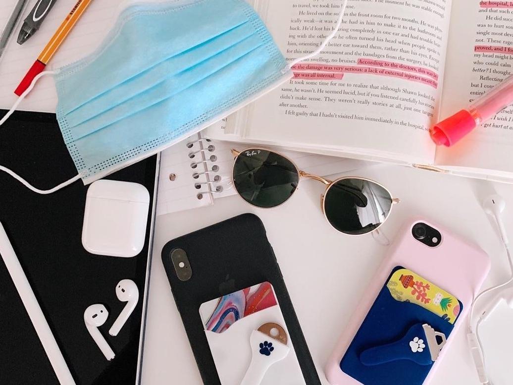 The Keyper phone wallet secures a student's ID card and key for easy access. The product is laid out with other student necessities, including a face mask, ear buds, sunglasses, a textbook and highlighters.