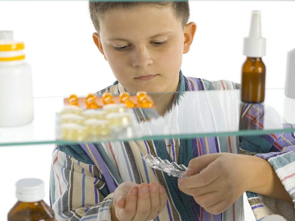 A child is seen through a medicine cabinet punching pills out into his hand. In the foreground, the cabinet contains several different medications.