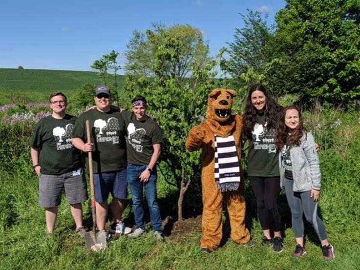 A row of men and women in matching T-shirts stand in tall grass with someone dressed as the Penn State Nittany Lion. One man holds a shovel.