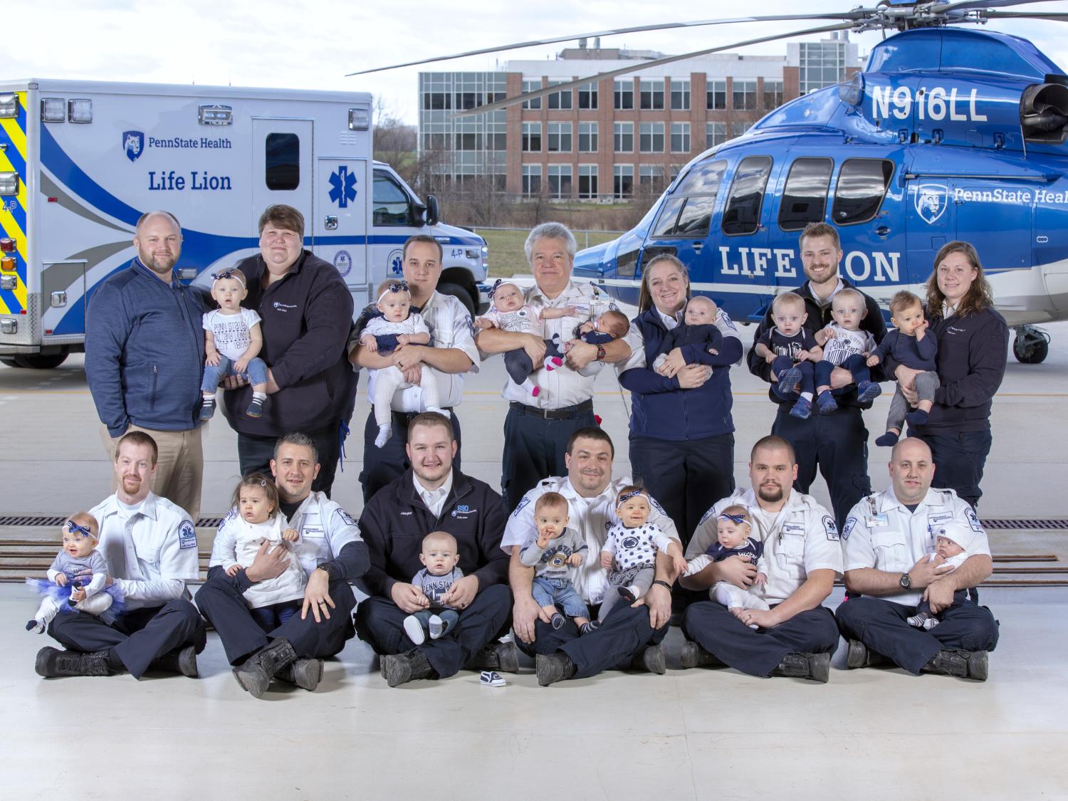 Thirteen adults pose for a photo, holding a total of 15 one-year-old children. In the near background is a Life Lion ambulance and a Life Lion helicopter. In the distant background is a brick building with dark windows.