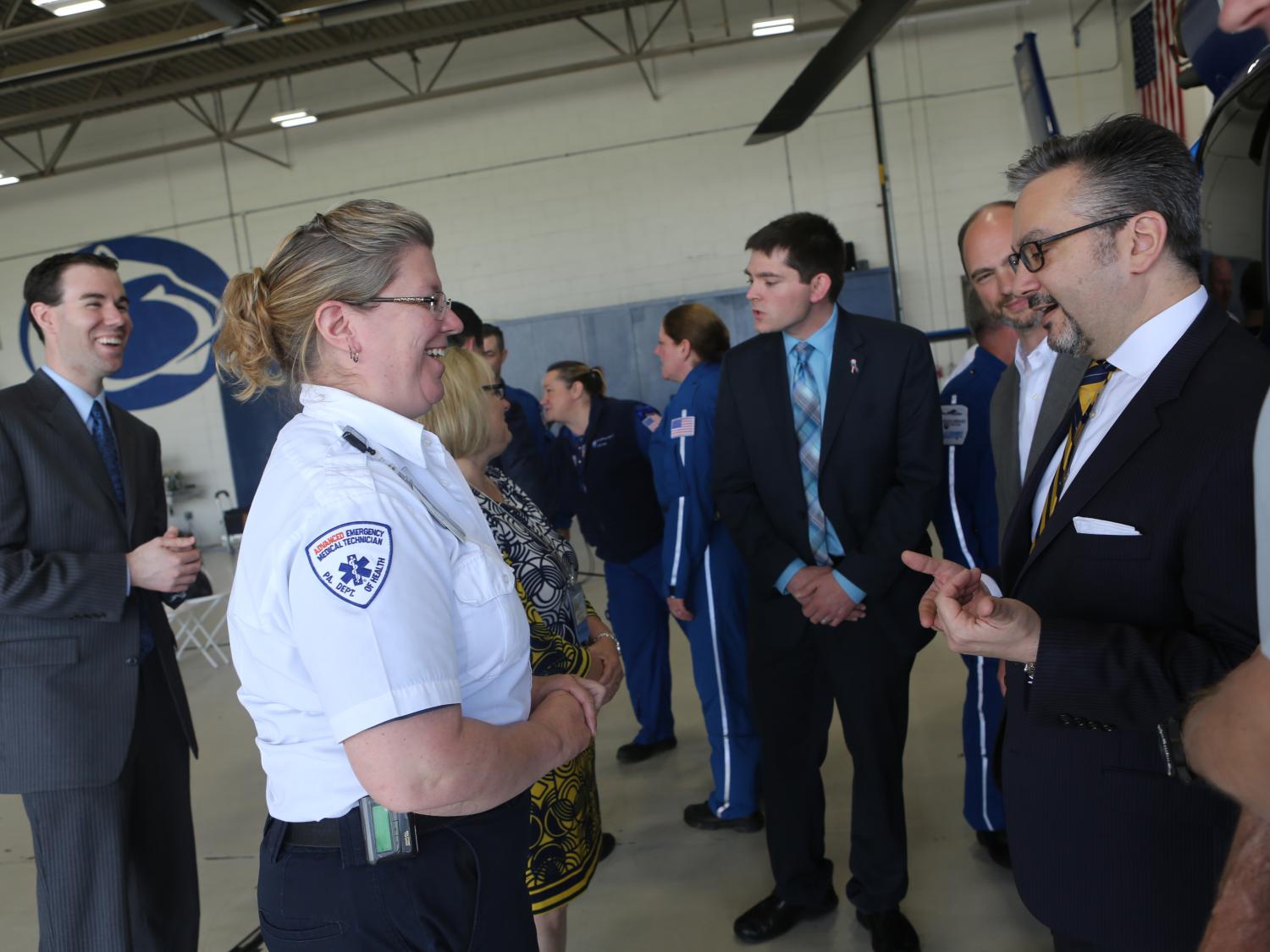 A woman in an EMS uniform speaks with a man in a suit. A helicopter is mostly off-camera, to the right. Other people are in the area – a helicopter hangar – standing and talking.