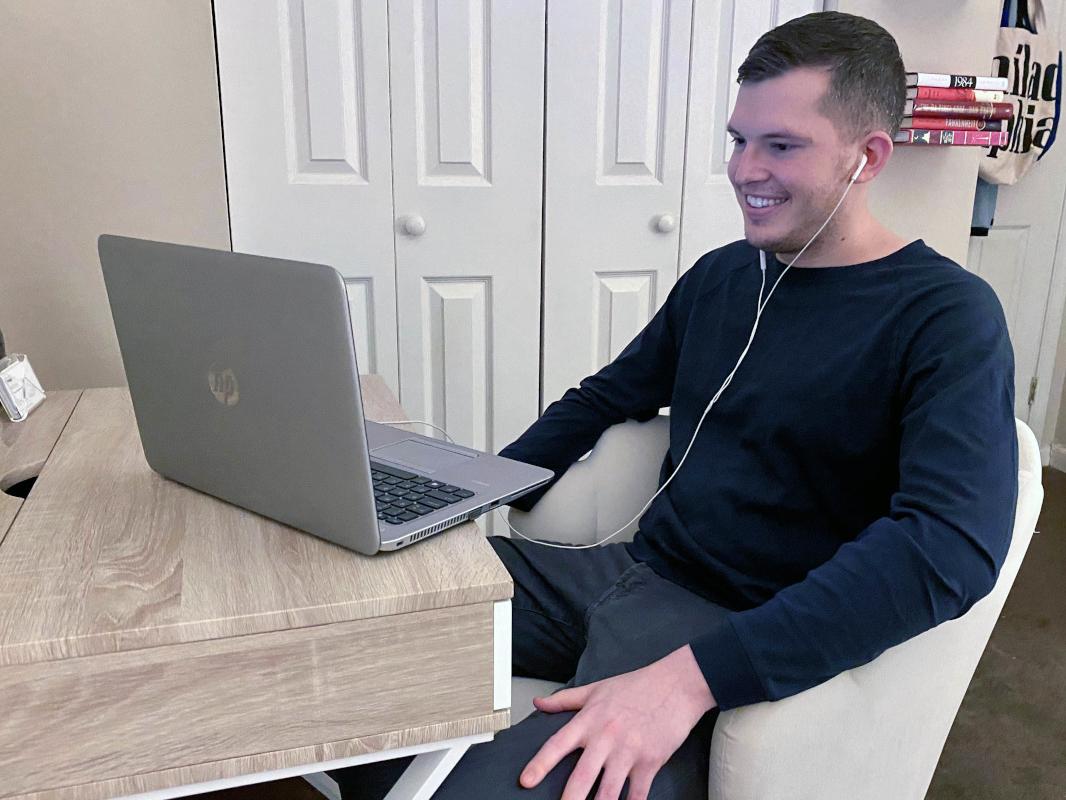 A man sits at a desk, wearing earbuds, looking at a laptop screen. He smiles slightly. A door, bookshelf and closet are in the background.