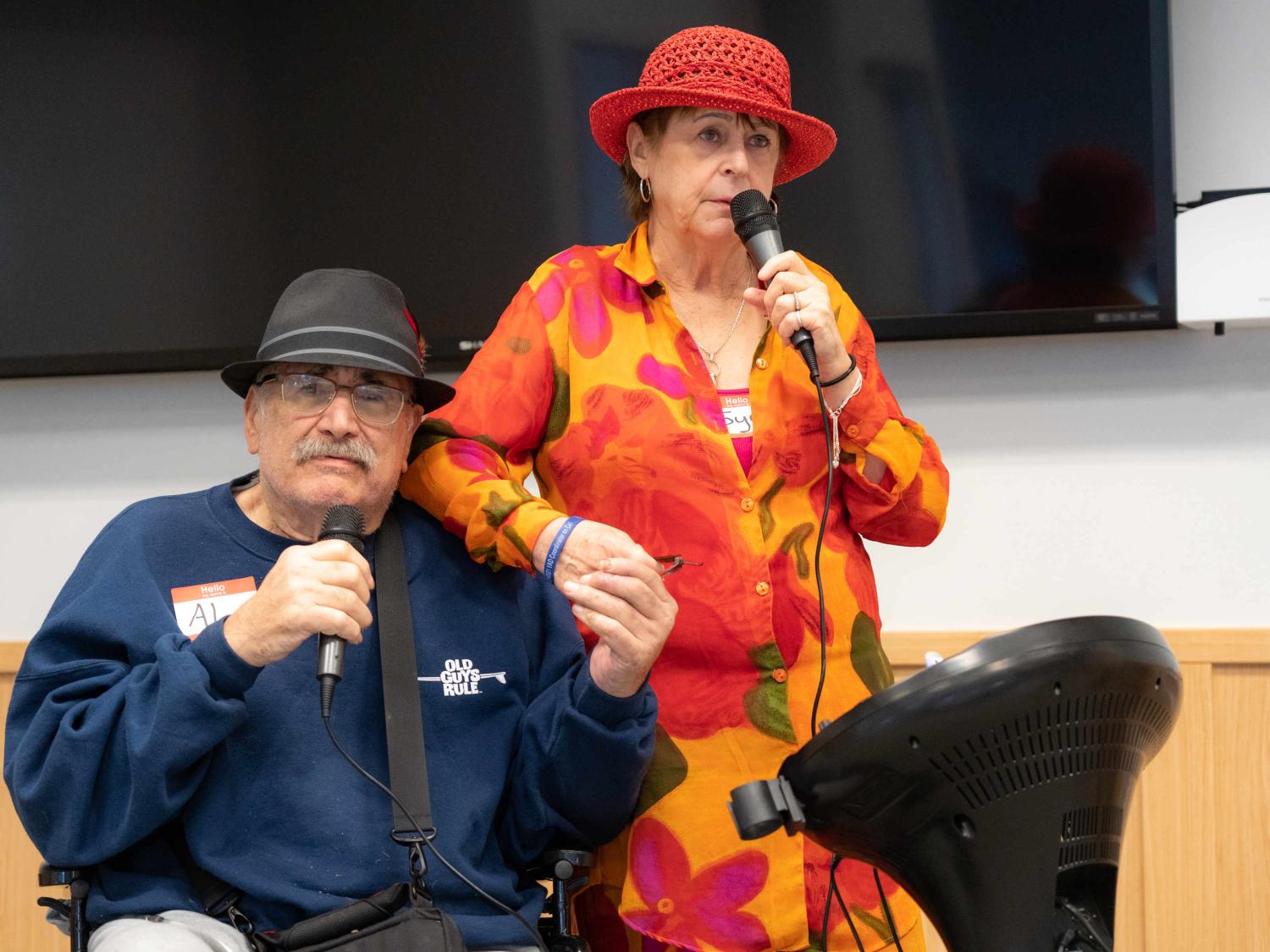 Al Dolatoski and his wife Joyce hold hands and hold microphones as they sing Sonny and Cher’s “I Got You Babe” during the LVAD Celebration of Life. Al is sitting in a wheelchair and has an LVAD hanging by a strap around his shoulder.