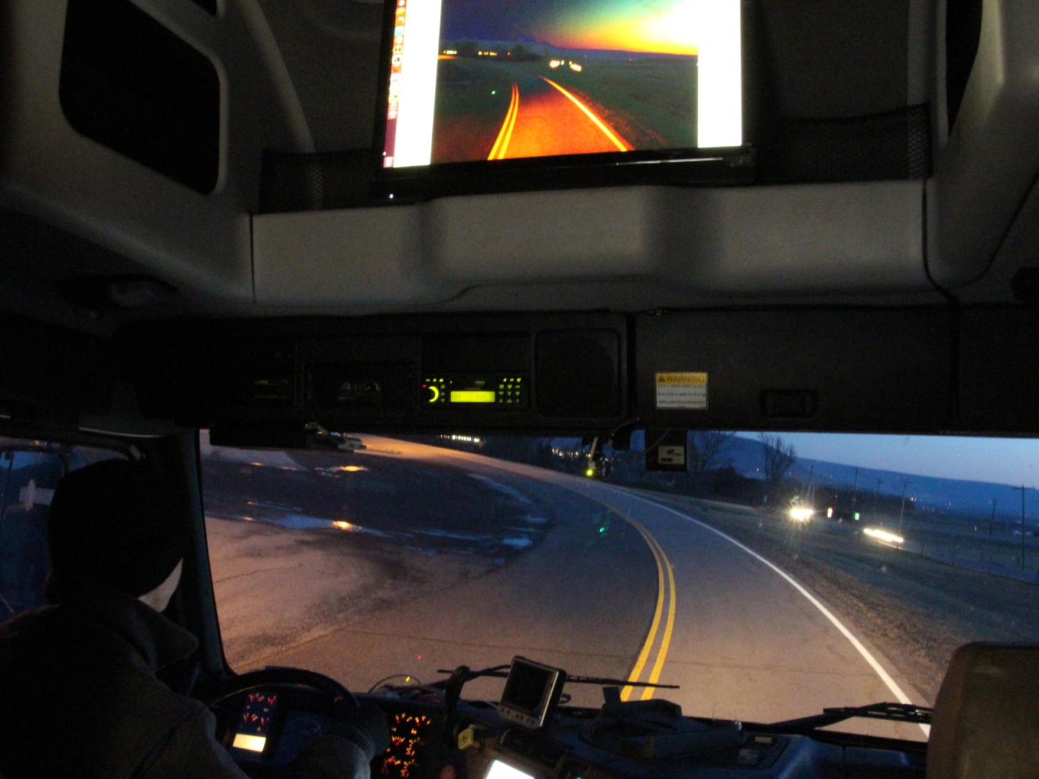 View of a marked roadway from inside a truck cabin with computer screens showing an image of the same road.