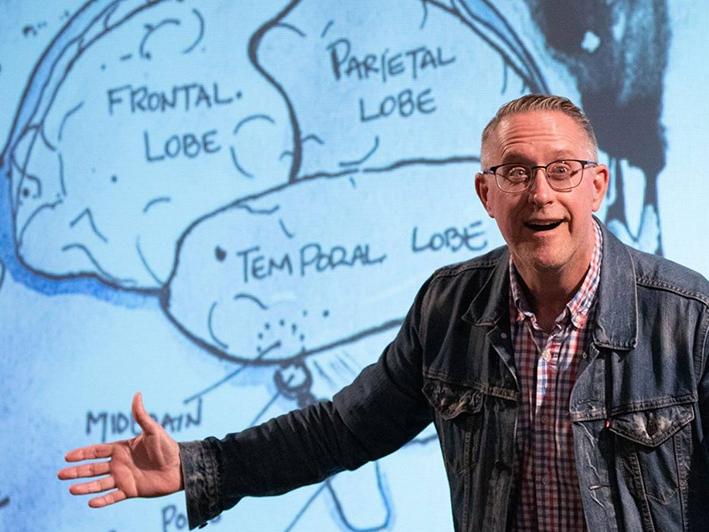 The professor gestures as he stands in front of the projected image of a hand-drawn brain.