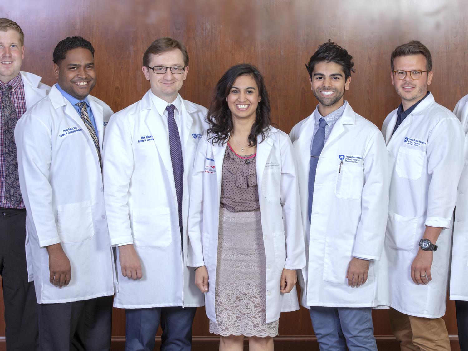 Six men and one woman wearing lab coats with the Hershey Medical Center logo and their names on them stand in a row and smile. The men are wearing shirts and ties, and the woman is wearing a skirt and top. The wall behind them is made of wood paneling.