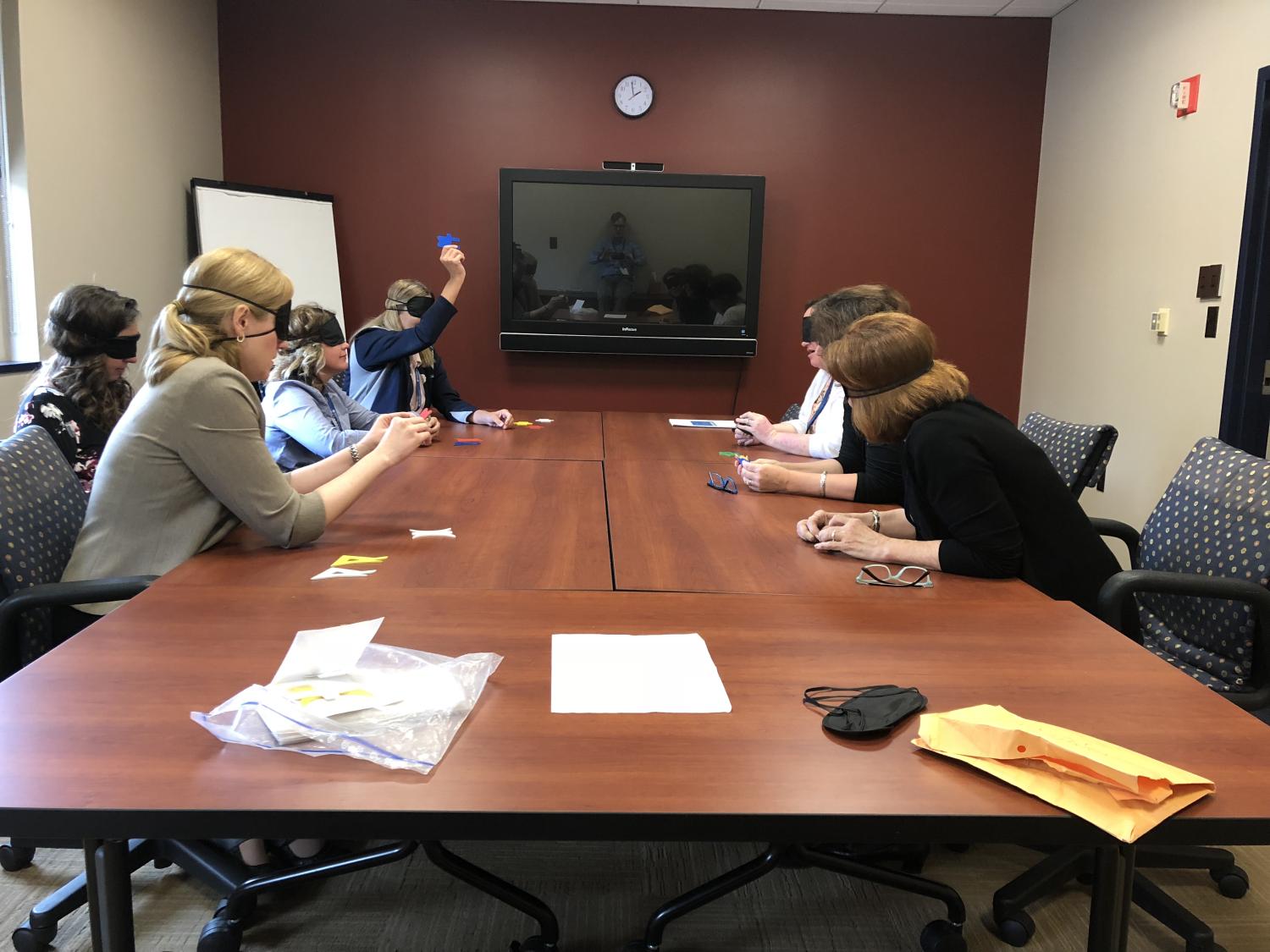 Seven people sit at a conference room table. Each wears a blindfold and has several cards in front of them on the table. One woman is holding a blue card in the air.