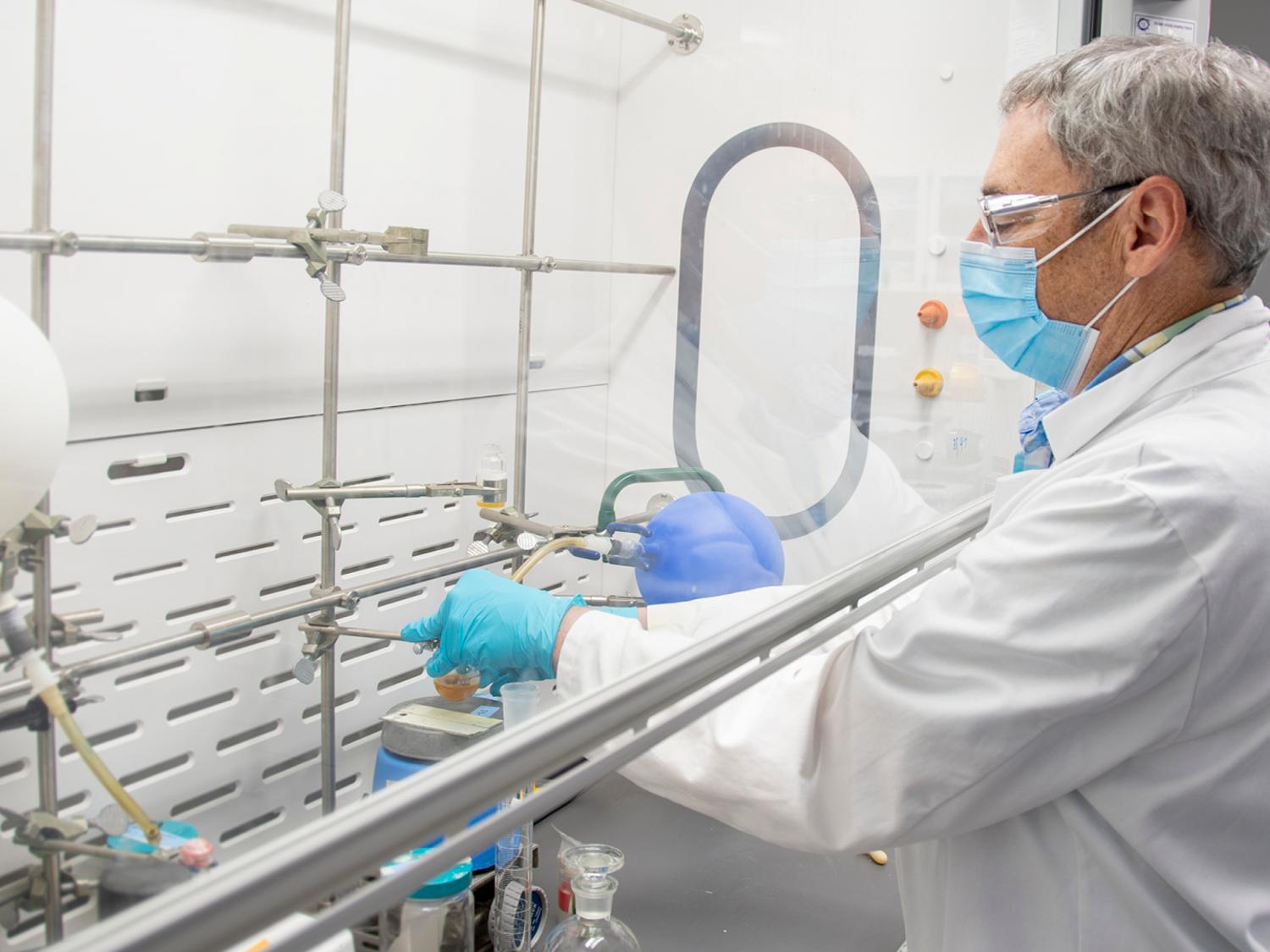 Man wearing lab coat, latex gloves, mask, and protective goggles pours a liquid into another canister in an organic chemistry lab