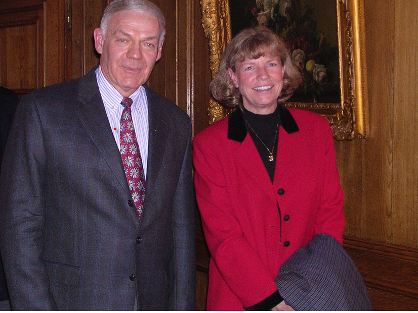 A man wearing a suit and tie and a woman in a red blazer stand and smile at the camera. 