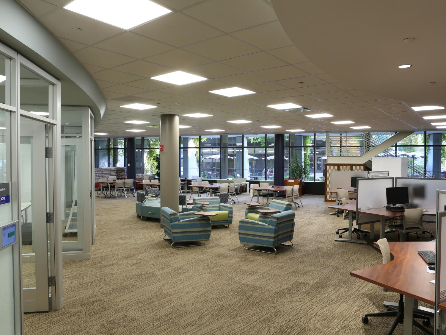 Tables and chairs with modern designs are seen placed across the room. Computer work station desks are to the right; glass-enclosed meeting and work room space is to the left.