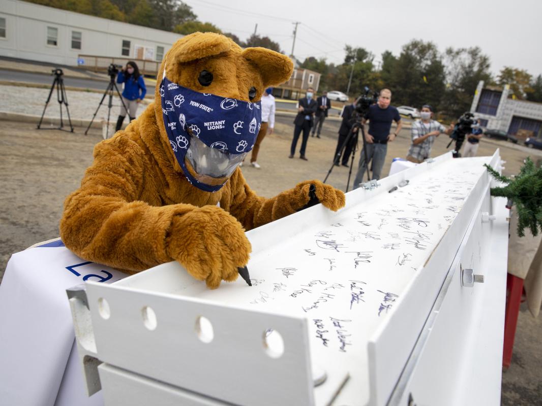 The "Nittany Lion," wearing a facemask, uses a marker to sign a metal beam. Several people walk around in the background.