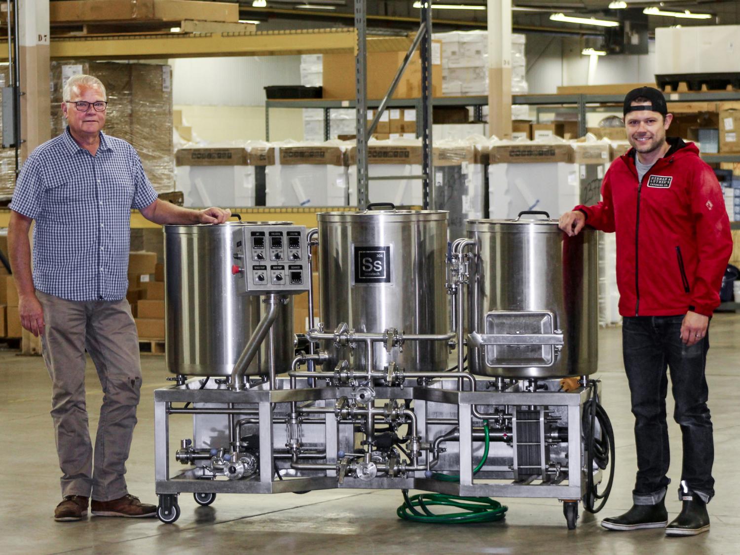 Two men stand next to a professional-grade brewing system
