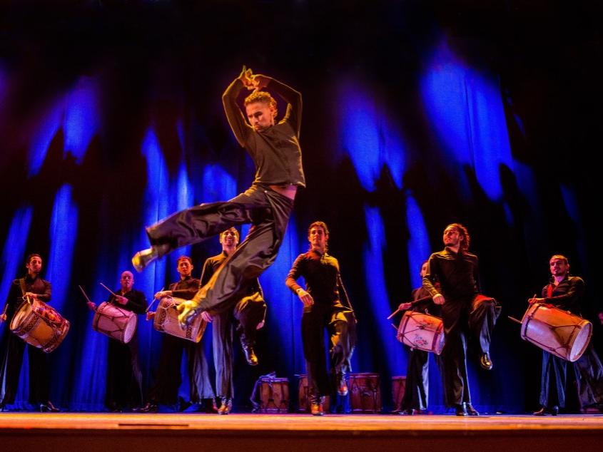 A dancer holds his hands straight above his head and kicks both legs out to the side while a line of dancers and drummers stand behind him and watch.