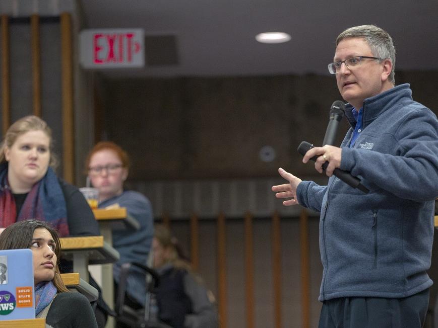 Paul Haidet speaks into a microphone at the rear of a classroom. Students type on laptops or look at the large screens and chalk board in the front of the room. A row of five students are seated, facing the class in the front of the room.