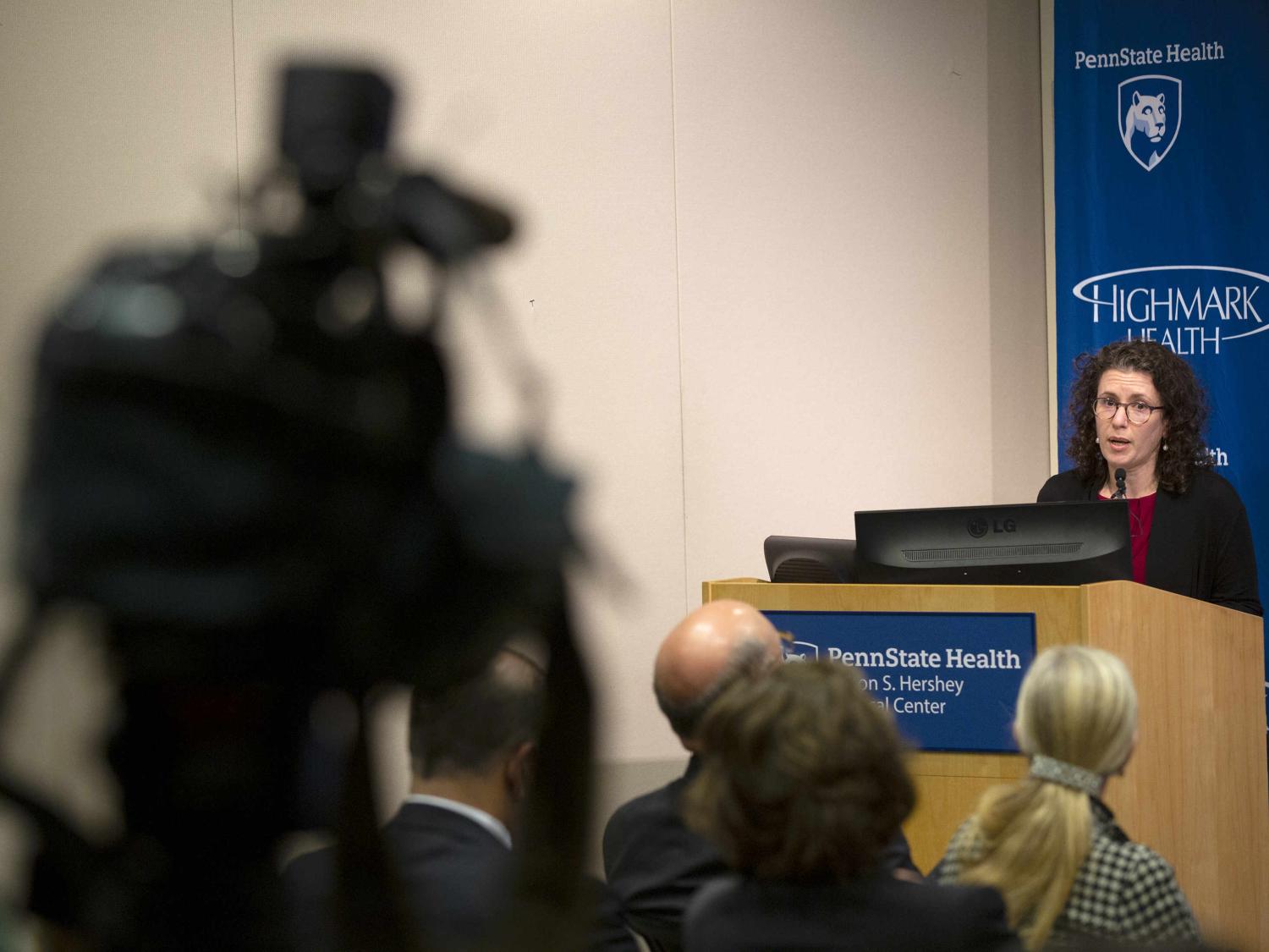 A woman stands at a lectern, speaking. People sit in chairs in the audience. A TV camera is to the left, in the foreground.