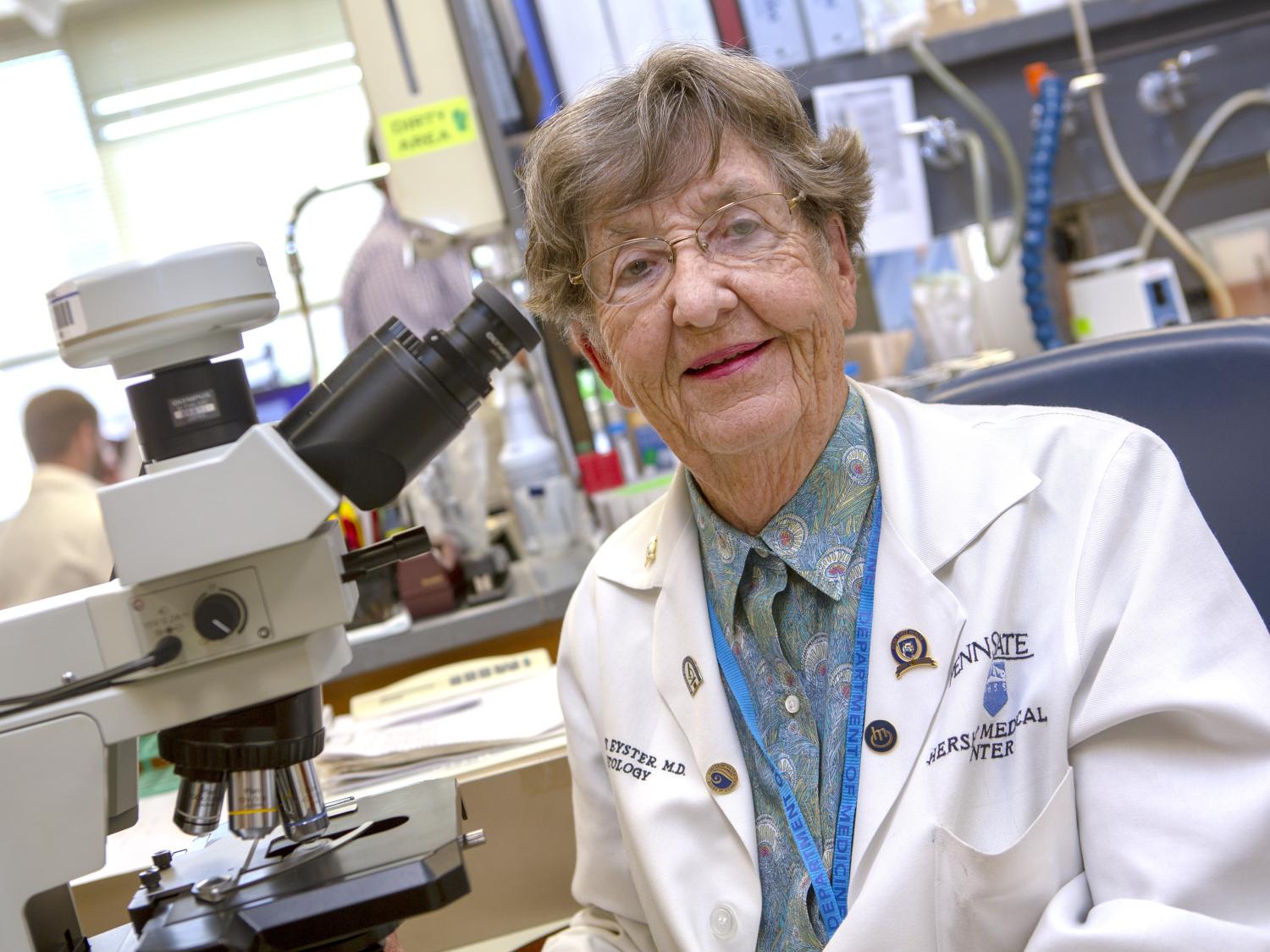 Dr. Elaine Eyster poses for a photograph in a lab, wearing a lab coat. A microscope is nearby.