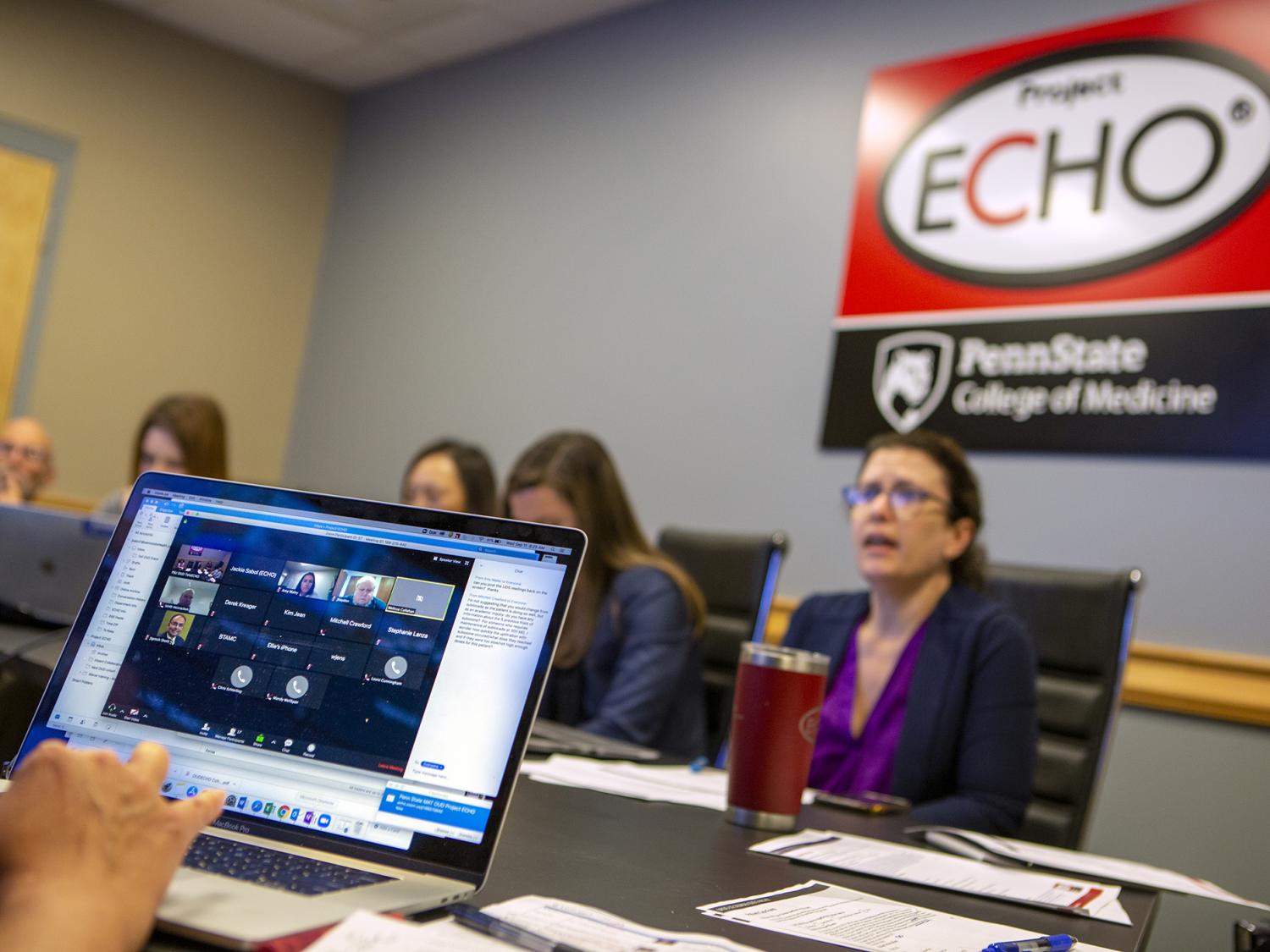 Several people sit around a conference table, on which there are papers, pens and computers. A sign on the wall bears the Project Echo and Penn State College of Medicine logos.