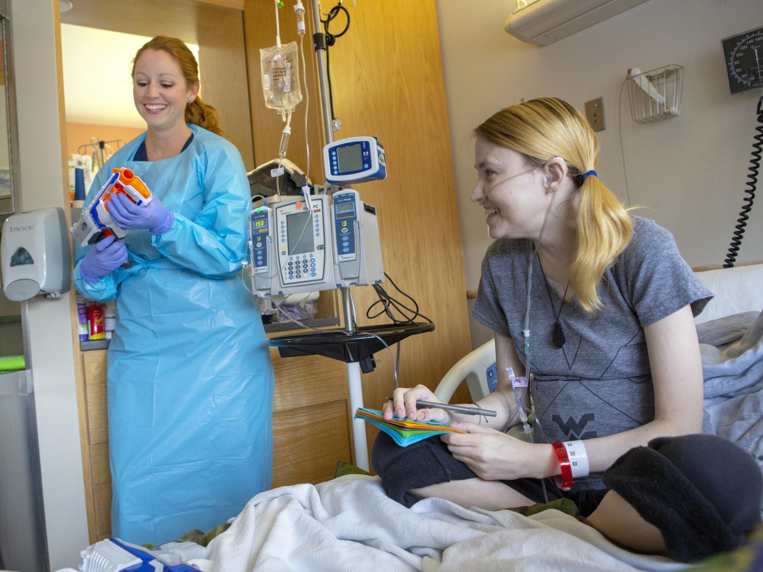 A woman sits up in a hospital bed, wearing a t-shirt and sweat pants. A nurse stands to the side of the bed, next to an IV pole, holding a Nerf dart gun. Both are smiling or laughing.