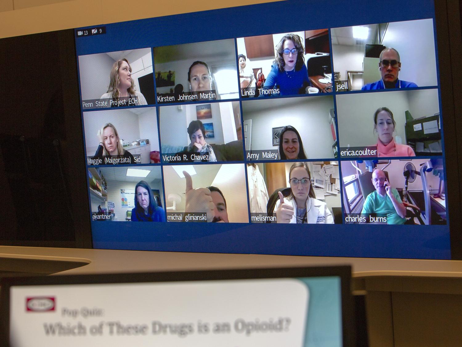 A large screen contains 12 mini screens showing physicians participating in Project ECHO. The participants are looking into their own web cams as they participate in the session. An out-of-focus screen reads “Pop Quiz: Which of these drugs is an opioid?”