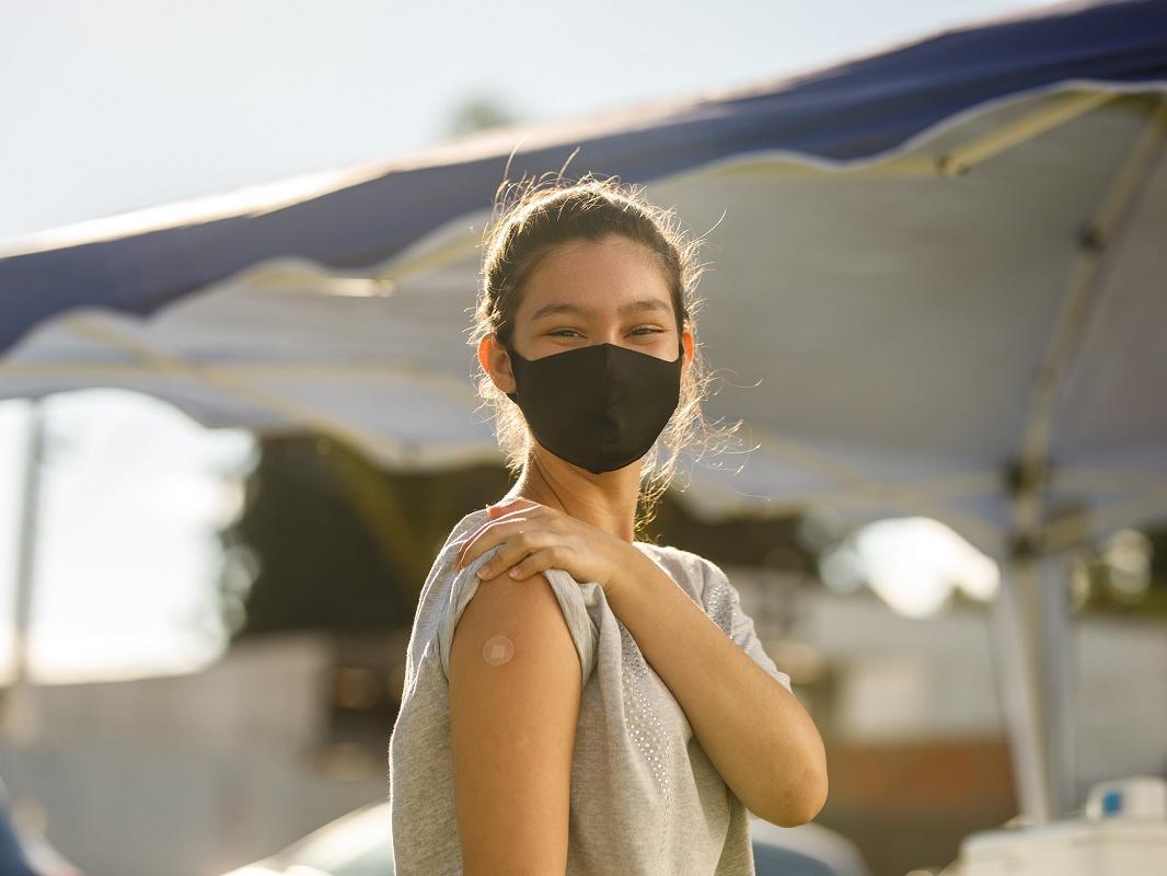 A girl in a mask shows the bandage where she received the vaccine on her arm.