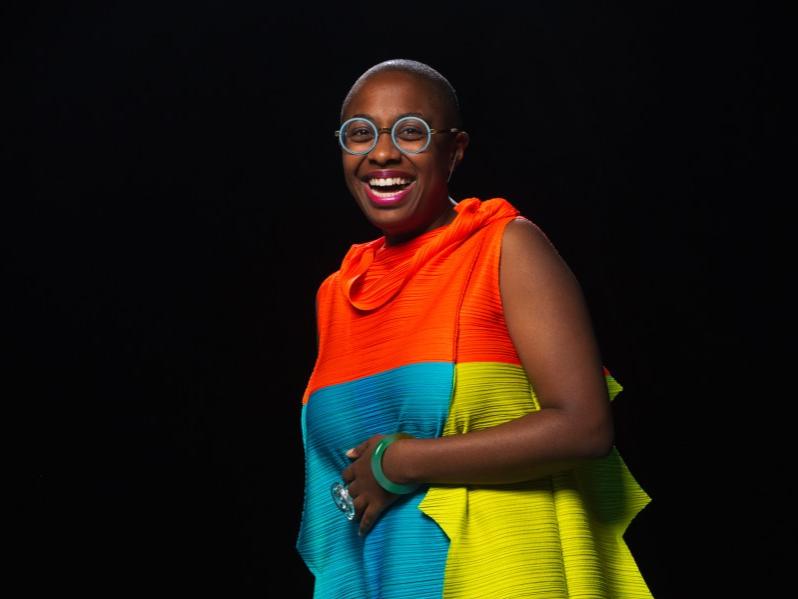 A woman smiles while she stands with her left arm wrapped around her waist.