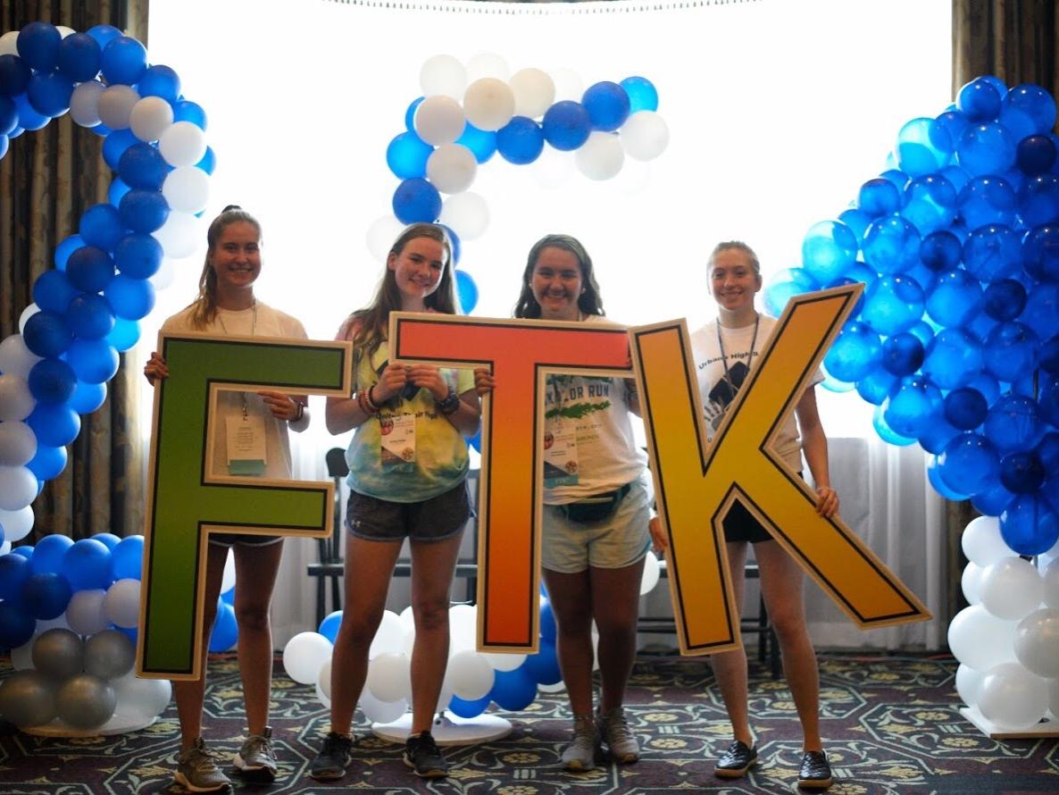 Four teenage girls hold oversized letters that spell out "FTK." Blue and white balloons and a window are in the background.