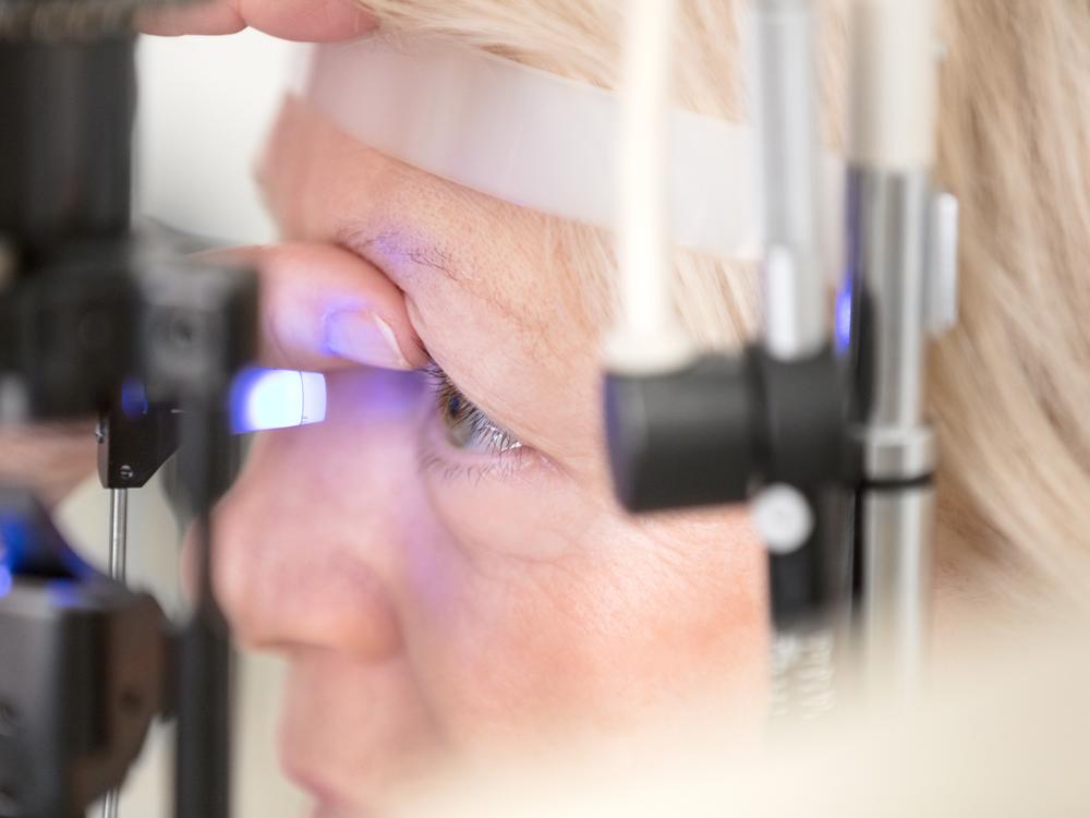 A close-up of a woman with her head placed against a piece of eye exam equipment. The equipment is shining a bright light into her eye, which is being held open by her thumb.