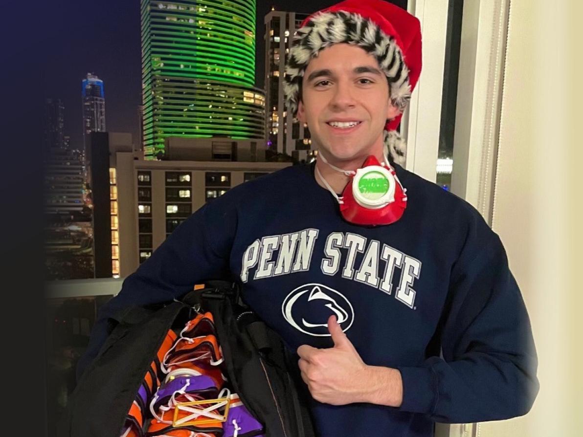 Marc Levine stands in front of a window at night wearing a Penn State T-shirt and Santa cap. He is giving the thumbs up and smiling. He has a face mask hanging from his neck and is holding a large bag with many face masks inside.