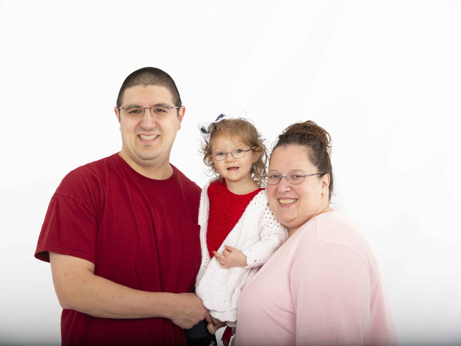 A young girl poses with her parents, all three of them smiling. The girl is being held up by her mom (at right) and dad (at left).