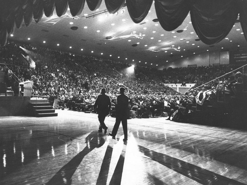 A dramatically lit, black and white photo of Martin Luther King Junior walking across the stage to the podium with a crowd of people in the audience